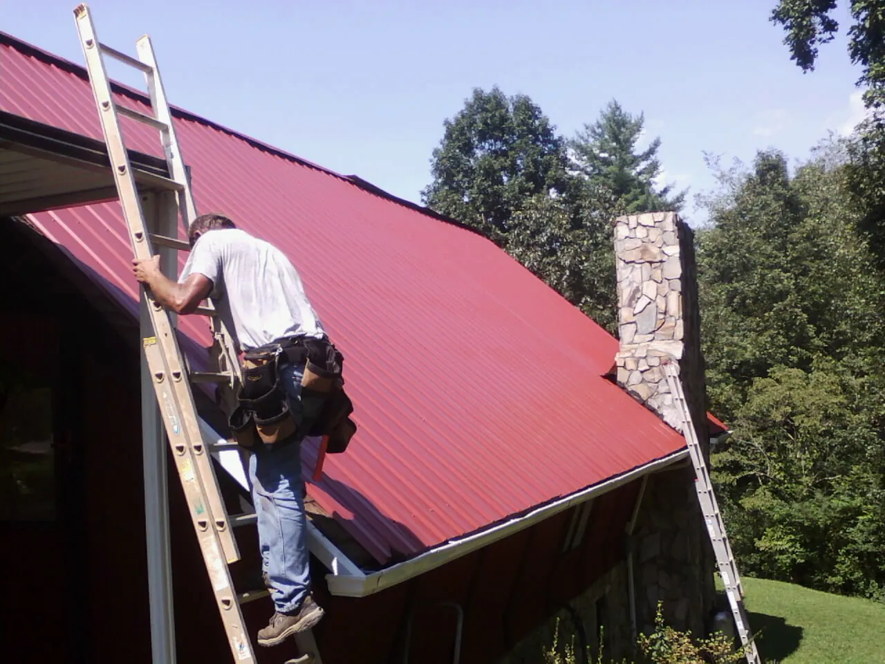 Roofer on a ladder beside a red metal roof and stone chimney — Soffit & Fascia Repair by Roofing Brothers in Avonmore